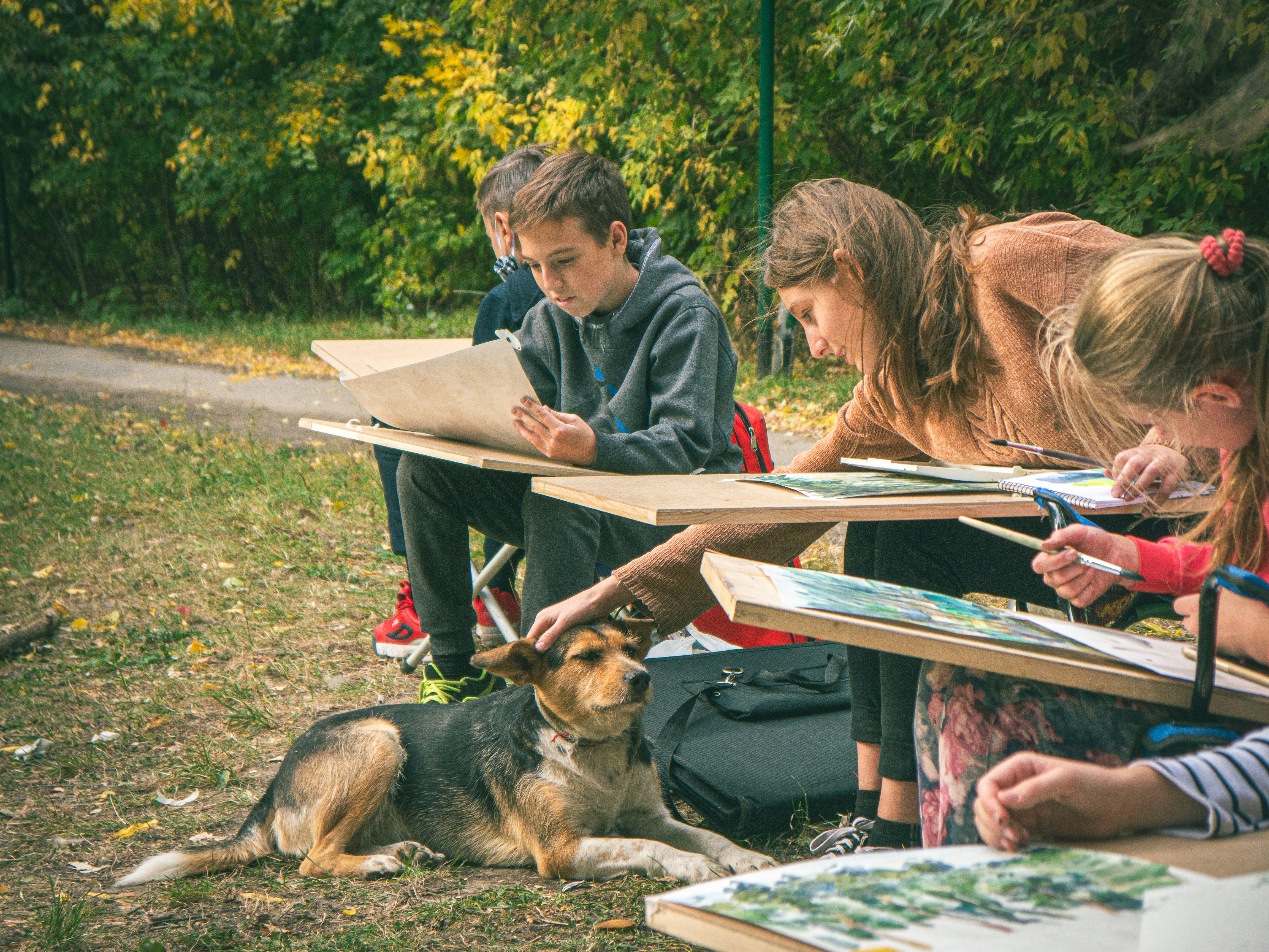 Art, Nature, and a Trusty Dog A candid moment from an outdoor art class where kids draw trees, a teacher gives guidance, and a loyal dog enjoys every minute of it. Captured with natural lighting in a 