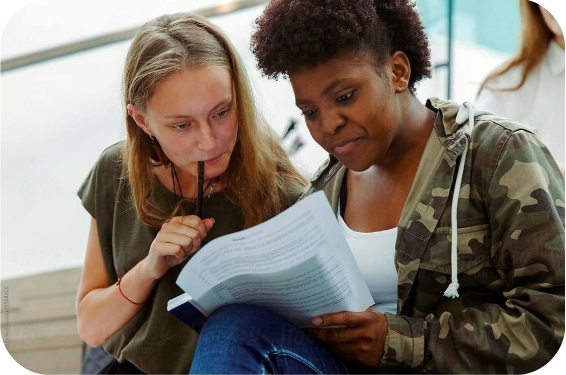 Two adult learners engaged in a study session, reviewing printed materials together. One holds a pen thoughtfully while the other turns the pages, highlighting collaborative learning in a supportive e
