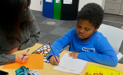 A young student writes on paper, focused on an educational activity, assisted by a tutor sitting beside him at a table with colorful learning materials.