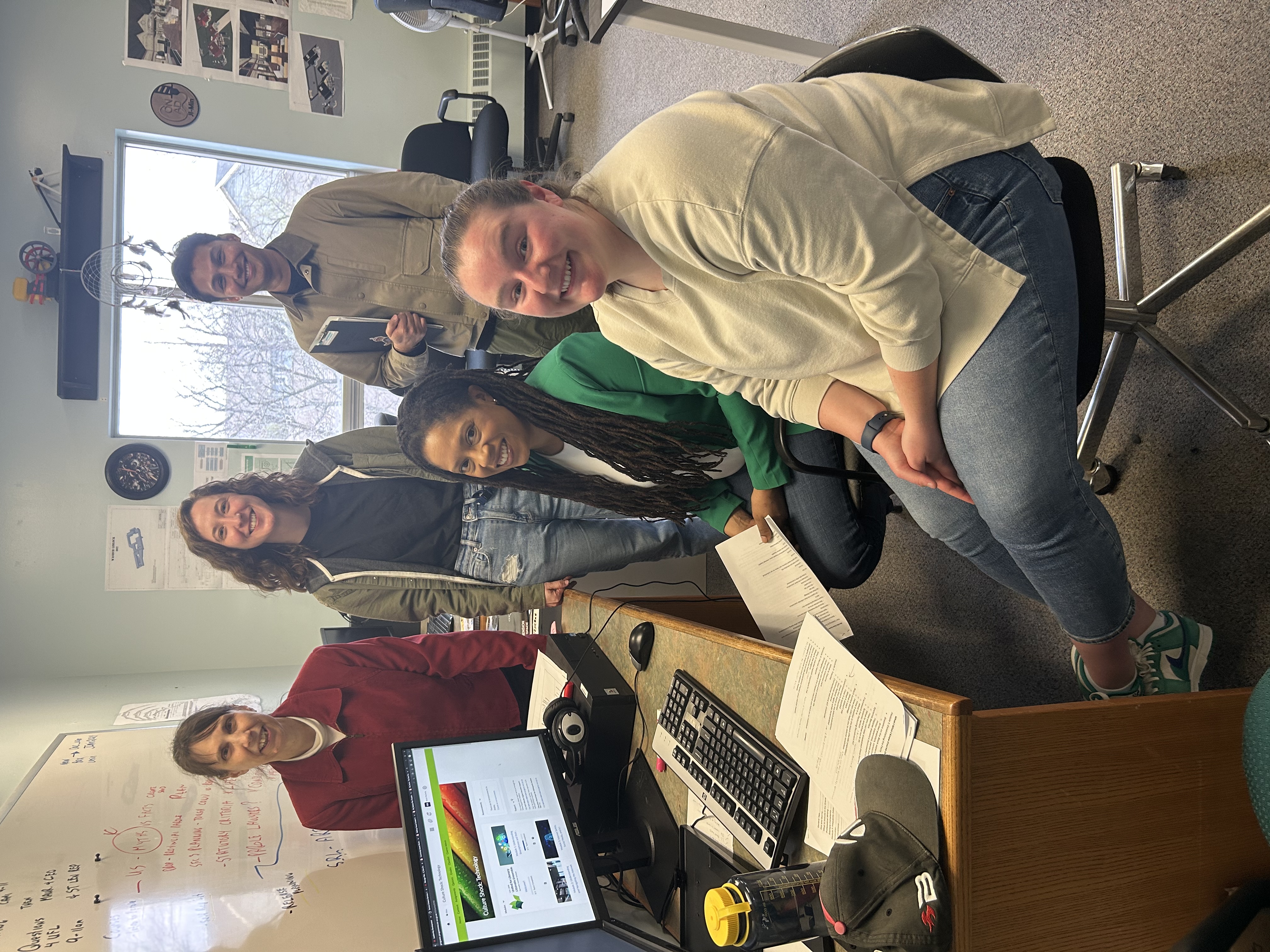 A group of five smiling adults gathered in an office around a desk with a computer. Two people are seated, while three stand behind them. Papers and a keyboard are visible on the desk. The computer sc