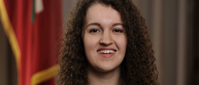 a woman with curly brown hair smiles in front of a flag