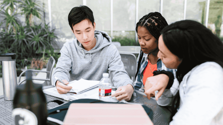 Three young adults studying together at a table, focused on a book they are reading collaboratively.