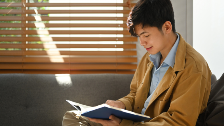 A young man sits by a window, sunlight falling across him as he tilts his head down with a slight smile, reading a book.