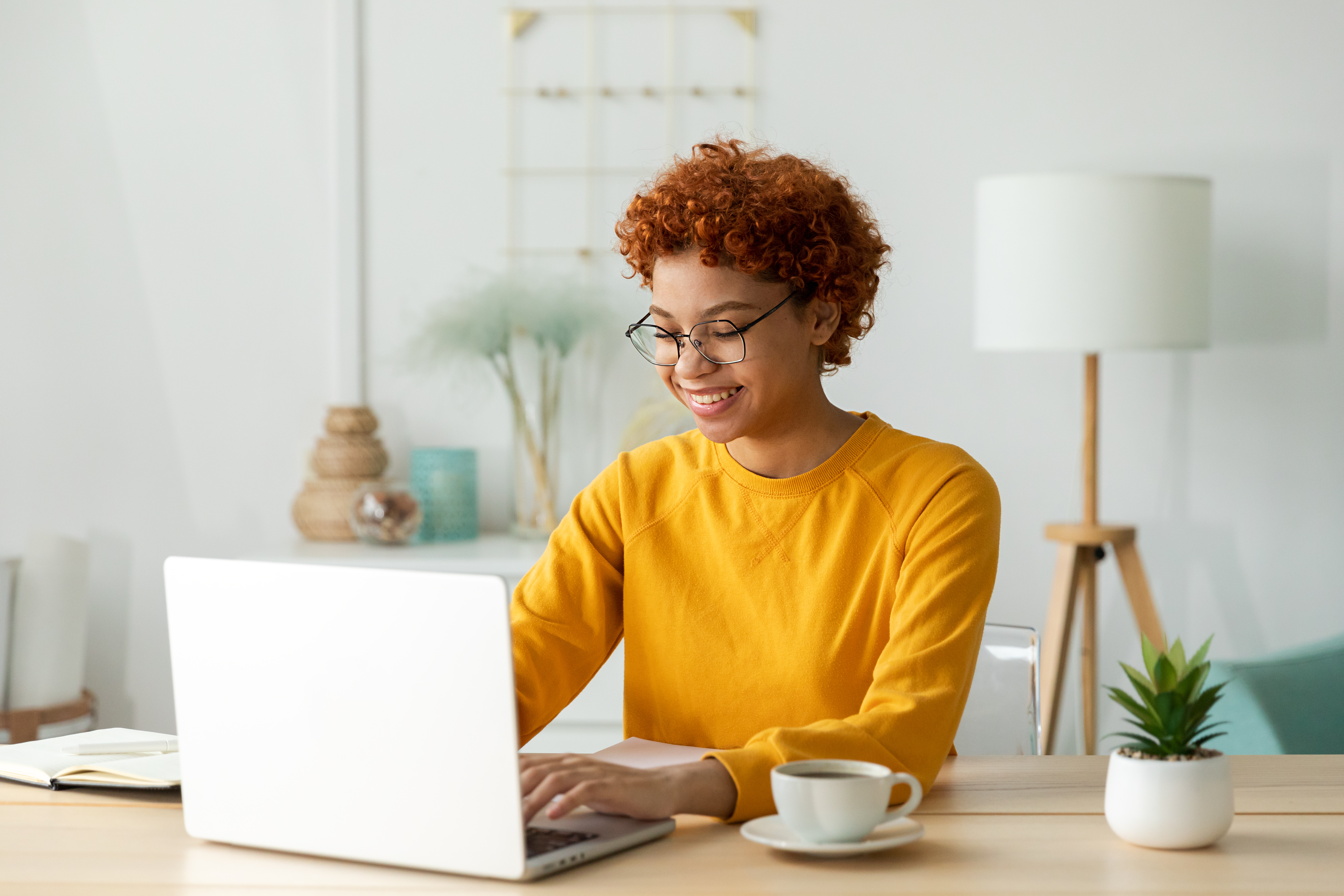 A black woman with a yellow sweater sitting at a desk, typing on her computer 