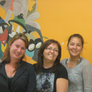 Three women stand in a row in front of an orange background.