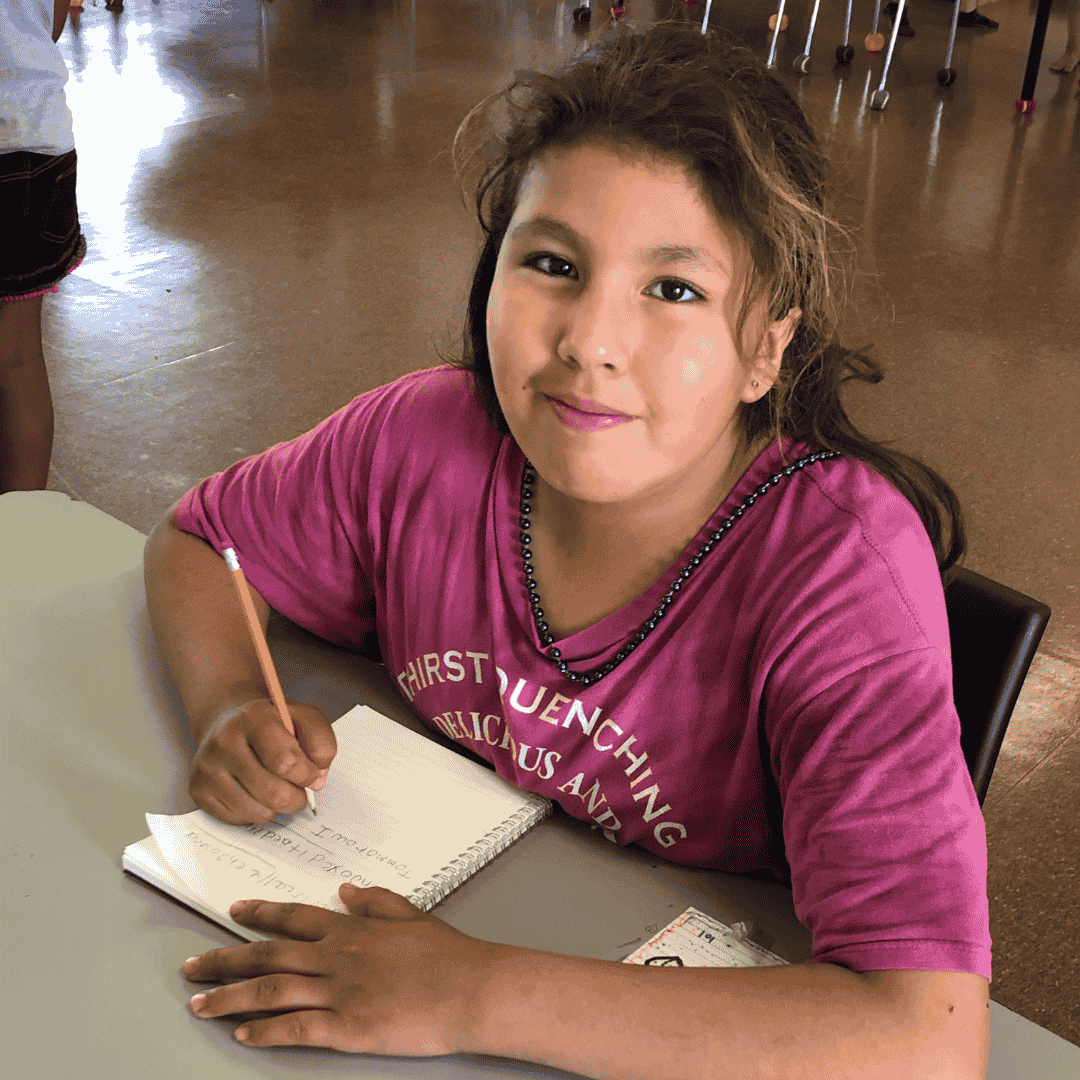 A young learner smiling at the camera and writing on a notebook